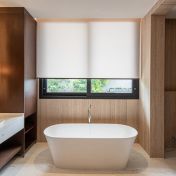 Sala de banho minimalista e sofisticada, apresentando uma persiana rolo em tecido técnico branco que garante privacidade total sem abdicar da luminosidade natural. O design do ambiente é marcado pelo equilíbrio entre o calor da marcenaria em madeira escura e a sobriedade das paredes em mármore travertino. A banheira de imersão freestanding e a bancada em mármore branco completam o cenário de refúgio particular, onde a persiana atua como um elemento essencial de conforto e purismo estético.