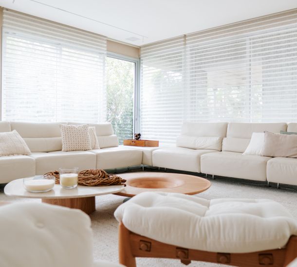 sala de estar moderna com amplas janelas cobertas por persianas translúcidas em camadas, permitindo a entrada suave de luz natural. O ambiente é composto por sofás em L de couro branco, uma mesa de centro de madeira e detalhes decorativos em tons neutros, criando uma atmosfera clean e aconchegante