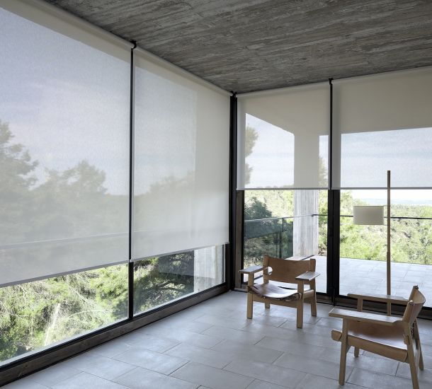 Sala com grandes janelas de vidro cobertas por cortinas sob medida em tom claro, permitindo a entrada suave de luz natural e vistas parciais para a área externa arborizada. O ambiente minimalista inclui duas cadeiras de madeira com design moderno e uma luminária de pé simples. O piso claro e o teto de concreto aparente dão um toque rústico e contemporâneo ao espaço, que se abre para uma varanda externa.