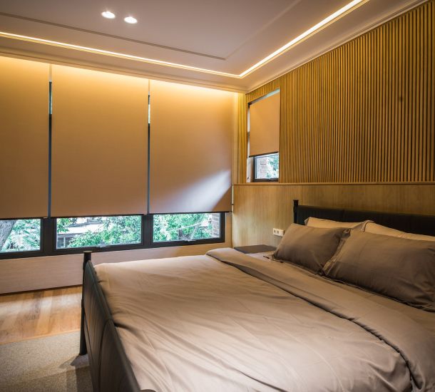 Quarto moderno com cortinas rolô fechadas cobrindo amplas janelas, criando um ambiente aconchegante e bem iluminado por luzes embutidas no teto. A cama de casal com roupas de cama em tons neutros complementa o design elegante, enquanto as paredes com painéis de madeira adicionam textura e sofisticação ao espaço