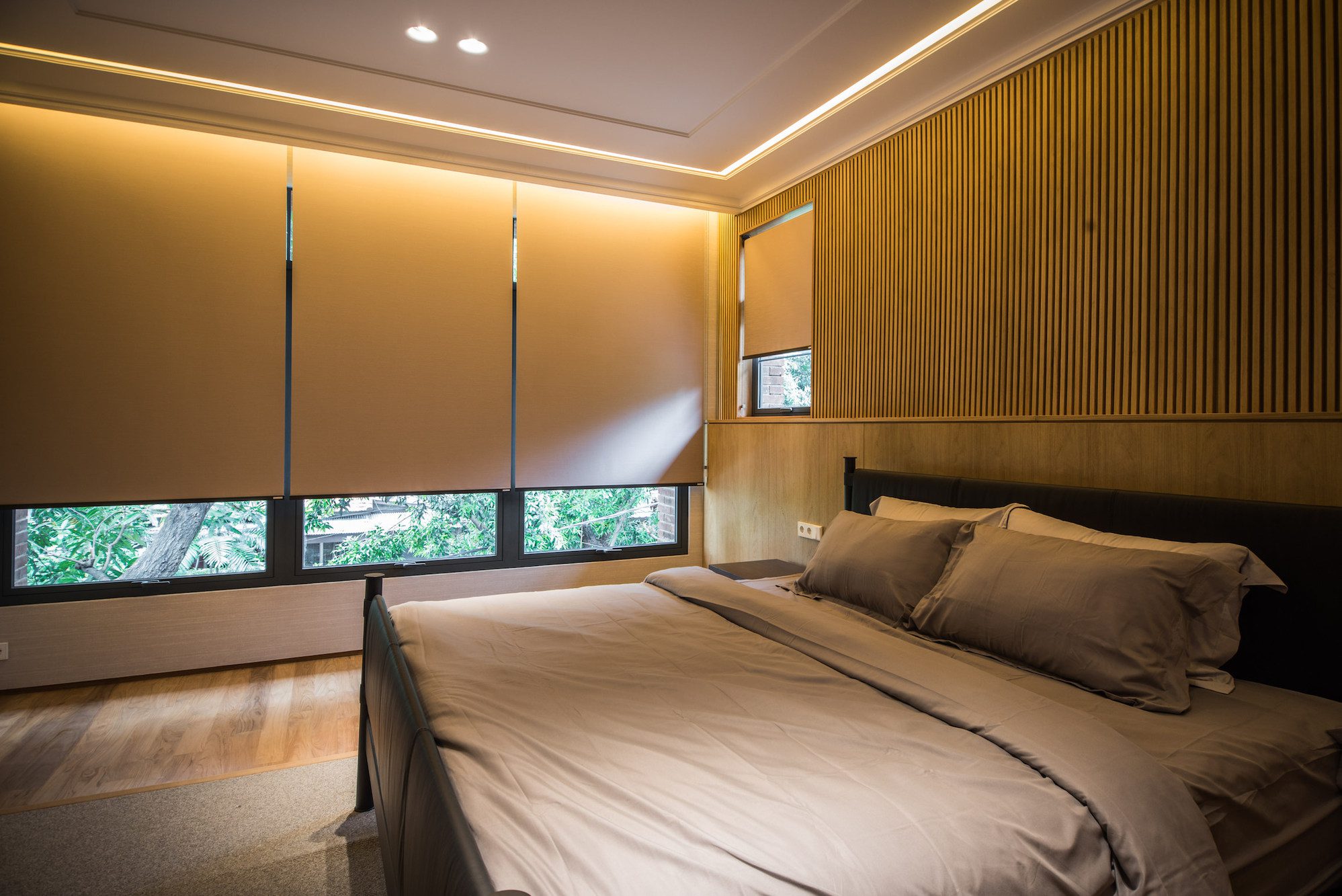 Quarto moderno com persianas automatizadas rolo bege fechadas, cama de casal em tons neutros e parede revestida em madeira.