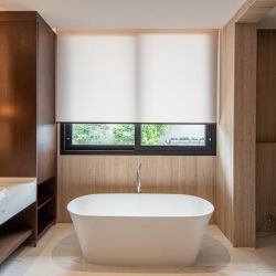Sala de banho minimalista e sofisticada, apresentando uma persiana rolo em tecido técnico branco que garante privacidade total sem abdicar da luminosidade natural. O design do ambiente é marcado pelo equilíbrio entre o calor da marcenaria em madeira escura e a sobriedade das paredes em mármore travertino. A banheira de imersão freestanding e a bancada em mármore branco completam o cenário de refúgio particular, onde a persiana atua como um elemento essencial de conforto e purismo estético.