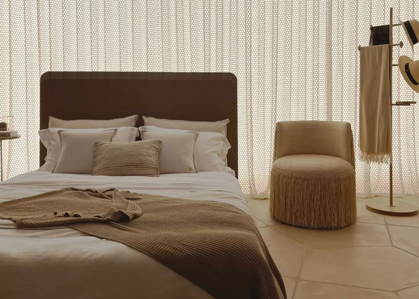 Quarto de casal sofisticado com uma ampla cortina de tela técnica hexagonal que cobre toda a parede de fundo, difundindo a luz de forma suave. A decoração segue uma paleta de cores neutras e terrosas, com cama posta, um pufe de franjas decorativo e um mancebo minimalista com chapéus.