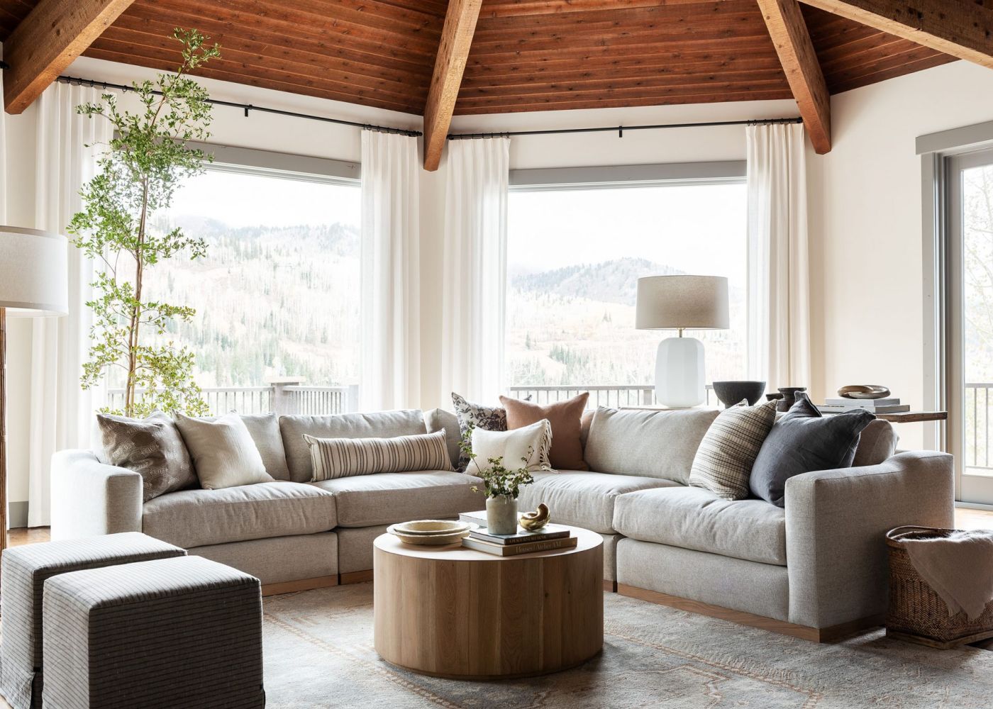 Living de alto padrão com arquitetura imponente e teto em madeira natural, onde cortinas de tecido fluido branco garantem leveza e emolduram a vista serena das colinas. A composição foca no conforto extremo, unindo um generoso sofá modular em tons neutros, texturas orgânicas nas almofadas e uma iluminação natural abundante que flui através das grandes janelas, transformando o espaço em um santuário de tranquilidade e convívio integrado à natureza.