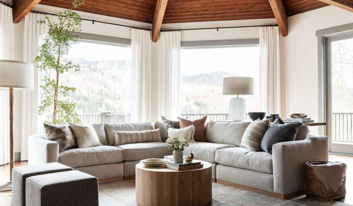 Living de alto padrão com arquitetura imponente e teto em madeira natural, onde cortinas de tecido fluido branco garantem leveza e emolduram a vista serena das colinas. A composição foca no conforto extremo, unindo um generoso sofá modular em tons neutros, texturas orgânicas nas almofadas e uma iluminação natural abundante que flui através das grandes janelas, transformando o espaço em um santuário de tranquilidade e convívio integrado à natureza.