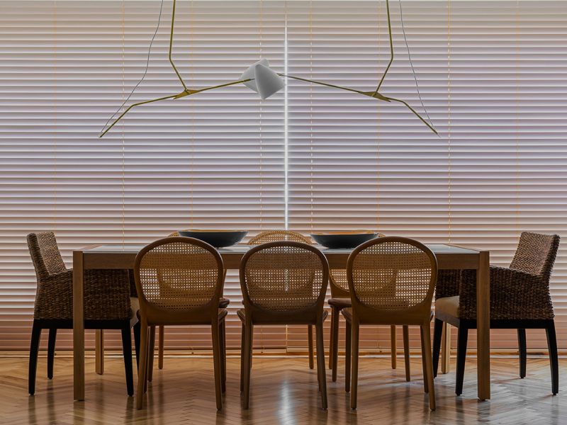 Sala de jantar refinada com persianas horizontais de design contemporâneo que proporcionam um controle milimétrico da luz, criando uma atmosfera acolhedora e privativa. A composição une o calor da madeira da mesa às texturas naturais das cadeiras em palhinha, sob a luz de um pendente escultural minimalista. O resultado é uma estética de luxo discreto, onde a geometria das persianas serve como moldura para um ambiente de convívio elegante e atemporal.