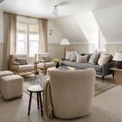 Sala de estar aconchegante com cortinas de tecido bege, poltronas de madeira clara, sofá cinza e mesa de centro redonda em fibras naturais.