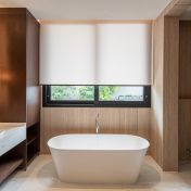 Sala de banho minimalista e sofisticada, apresentando uma persiana rolo em tecido técnico branco que garante privacidade total sem abdicar da luminosidade natural. O design do ambiente é marcado pelo equilíbrio entre o calor da marcenaria em madeira escura e a sobriedade das paredes em mármore travertino. A banheira de imersão freestanding e a bancada em mármore branco completam o cenário de refúgio particular, onde a persiana atua como um elemento essencial de conforto e purismo estético.