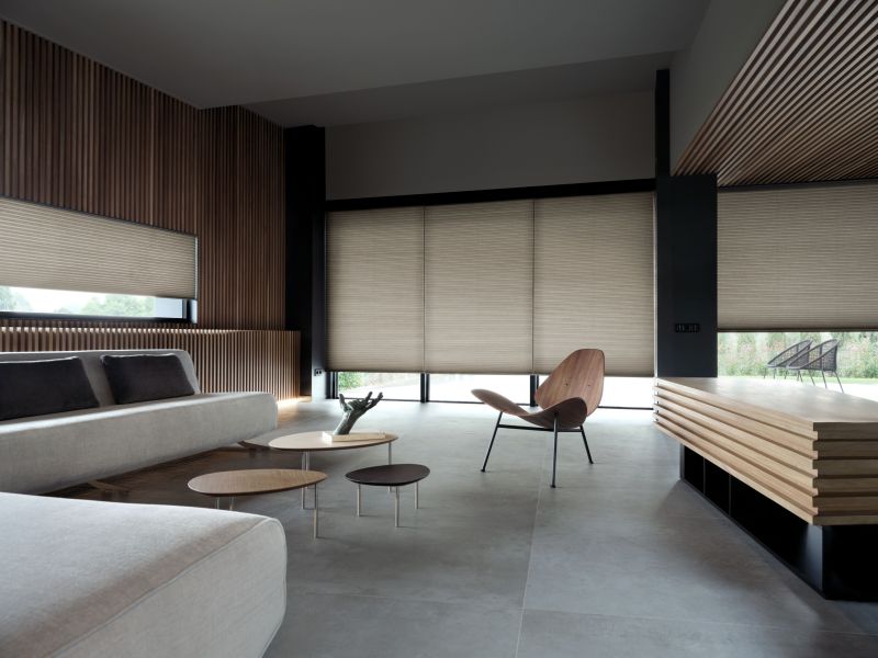 Sala de estar contemporânea e sofisticada, com amplas persianas celulares em tons neutros que proporcionam privacidade e conforto térmico. O design do ambiente é marcado pelo uso de madeira ripada nas paredes e teto, mobiliário minimalista de linhas puras — incluindo uma poltrona de design icônico e mesas de centro orgânicas — sobre um piso de porcelanato cinza, criando uma atmosfera de modernidade linear e serenidade.