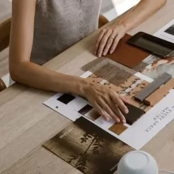 Vista superior detalhada de uma mesa de madeira clara onde mãos manuseiam uma seleção criteriosa de materiais para um projeto de design de interiores. Sobre a mesa, encontram-se amostras de tecidos texturizados, acabamentos em madeira, paletas de cores neutras e um catálogo com a frase "Make Every Space Matter". A cena personifica o suporte técnico e a curadoria de alto padrão oferecidos a arquitetos e clientes, focando na escolha precisa de texturas e materiais que conferem identidade e sofisticação a cada ambiente.